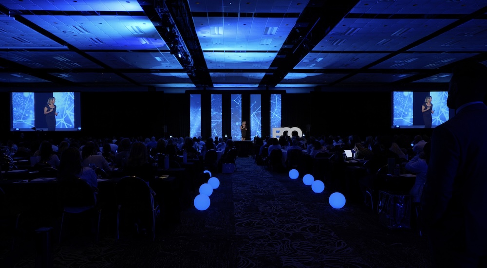Conference hall with a speaker on stage under blue lighting and large screens displaying visuals.