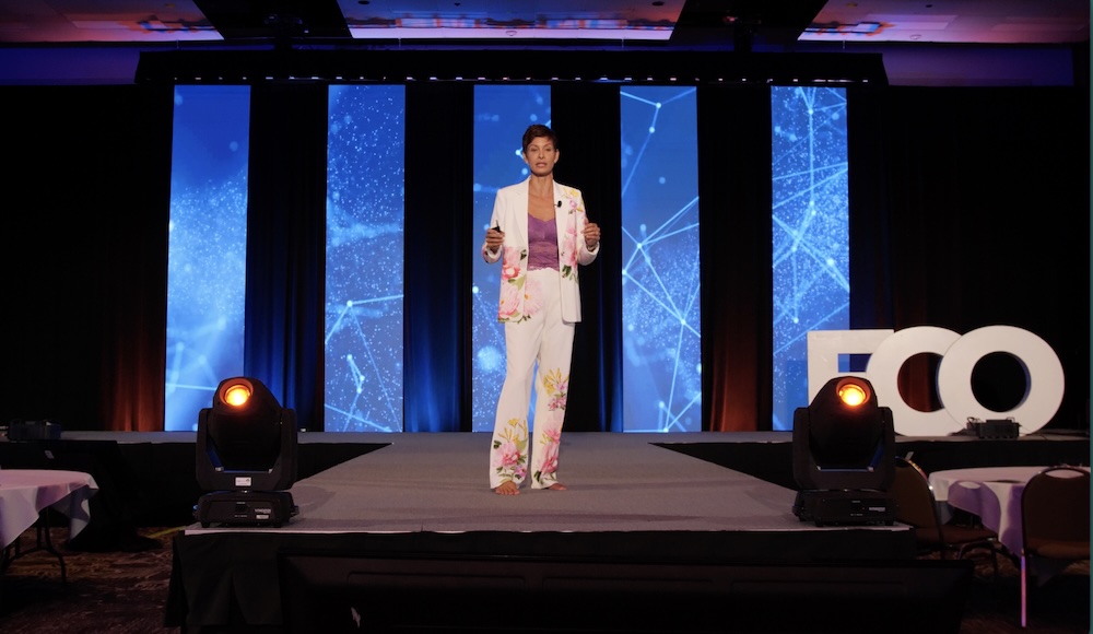 Female speaker on a stage wearing a white floral suit delivering a presentation against a blue, tech-inspired backdrop with vertical panels and light accents.