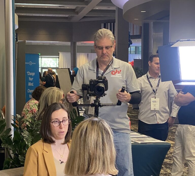 Man operating a camera on a stabilizer at a conference, filming two women seated in the foreground.