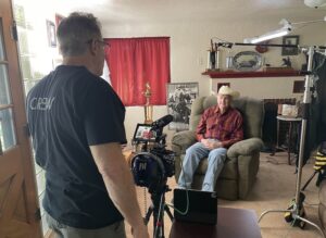 Crew member filming an elderly man seated in a living room for a video interview with camera on a tripod nearby