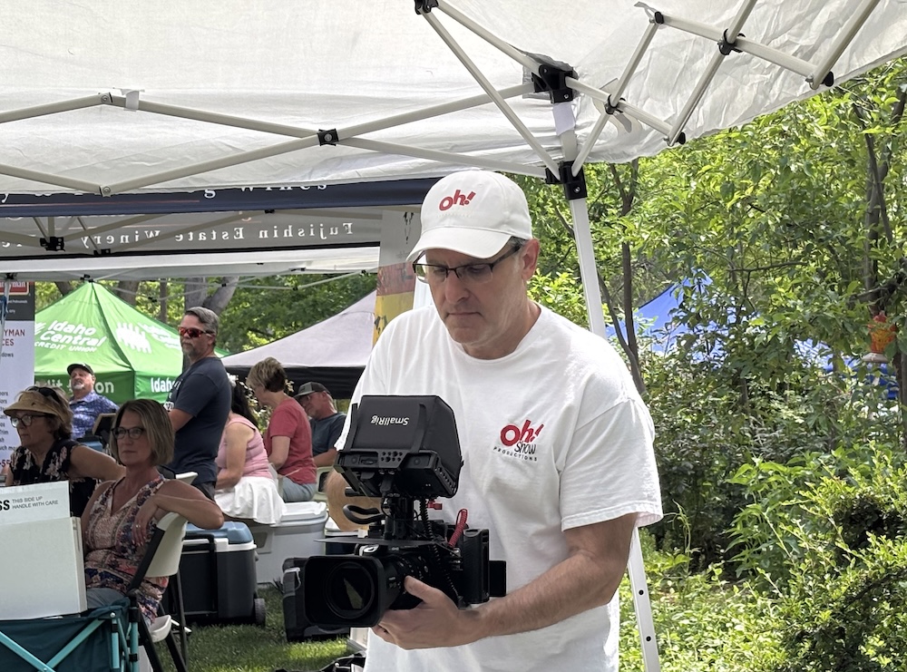 Videographer wearing a white oh! cap and shirt operates a large video camera under a white canopy at an outdoor event.