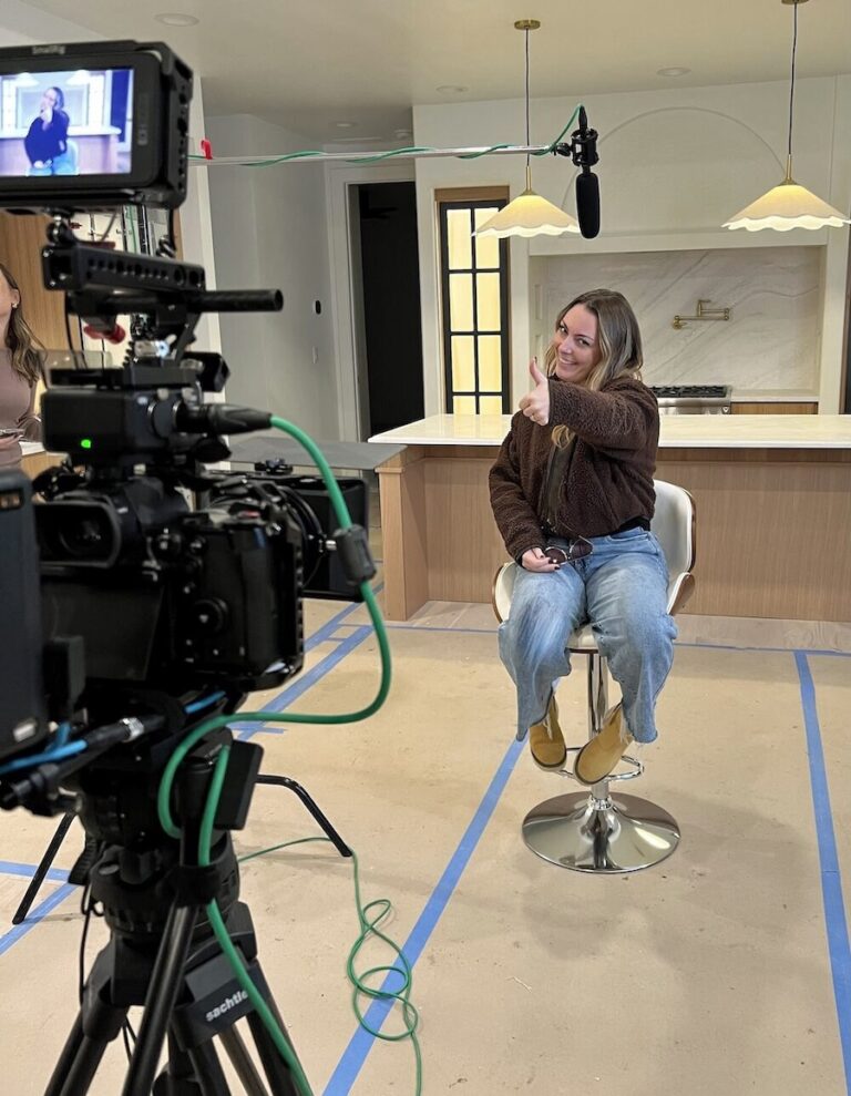 Woman in a brown jacket sits on a bar stool in a modern kitchen set, giving a thumbs-up, with a camera, mic boom, and monitor visible for a behind-the-scenes shoot.