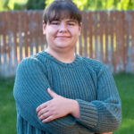 Person with short hair in a teal knit sweater stands outside with arms crossed, smiling. (outdoor portrait) LF