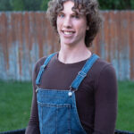 Smiling young man with curly hair wearing blue denim overalls and a brown long-sleeve shirt, outdoors in a yard with a wooden fence.